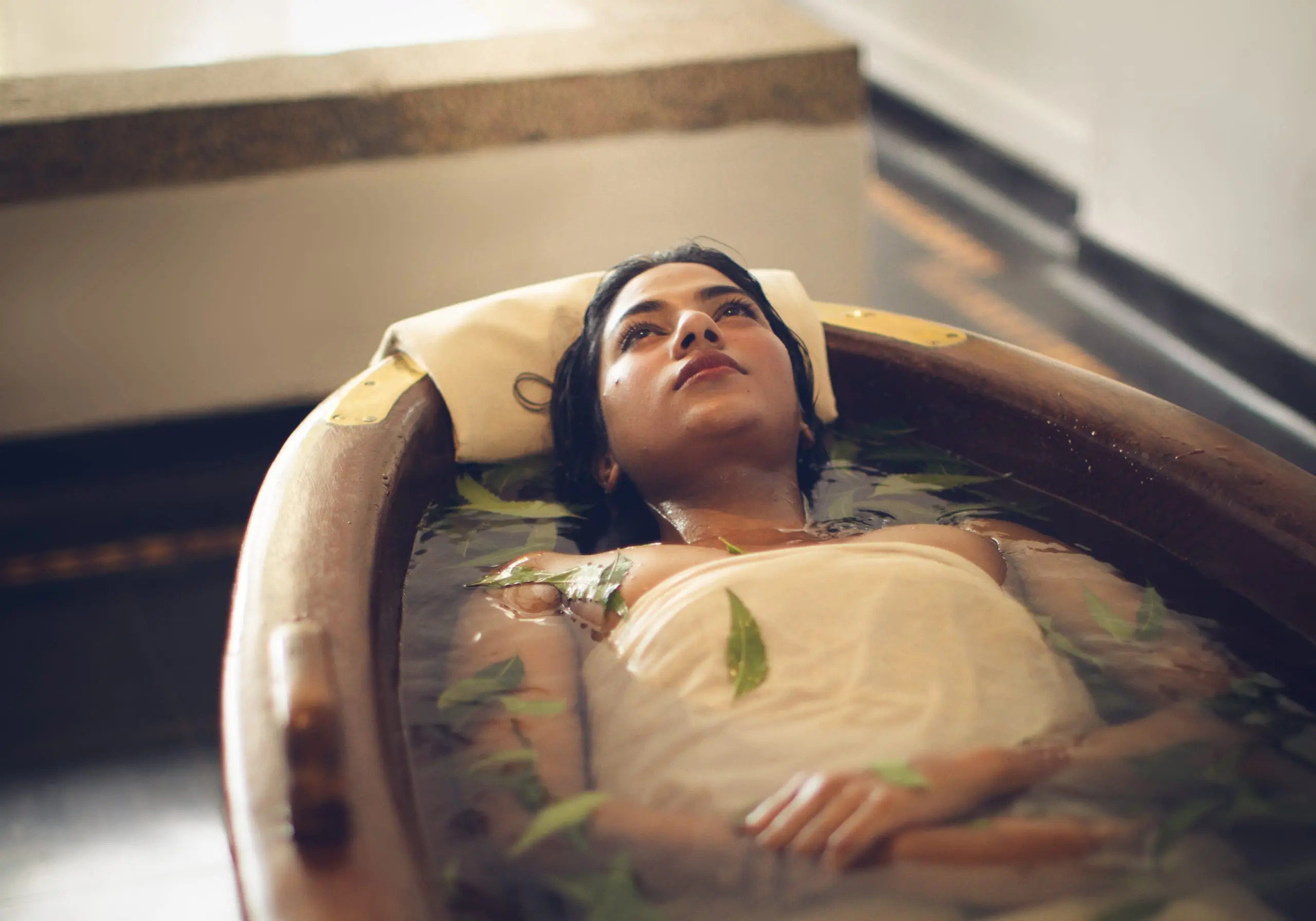 A guest rests in a wooden bath scattered with leaves, head on a cotton pillow.