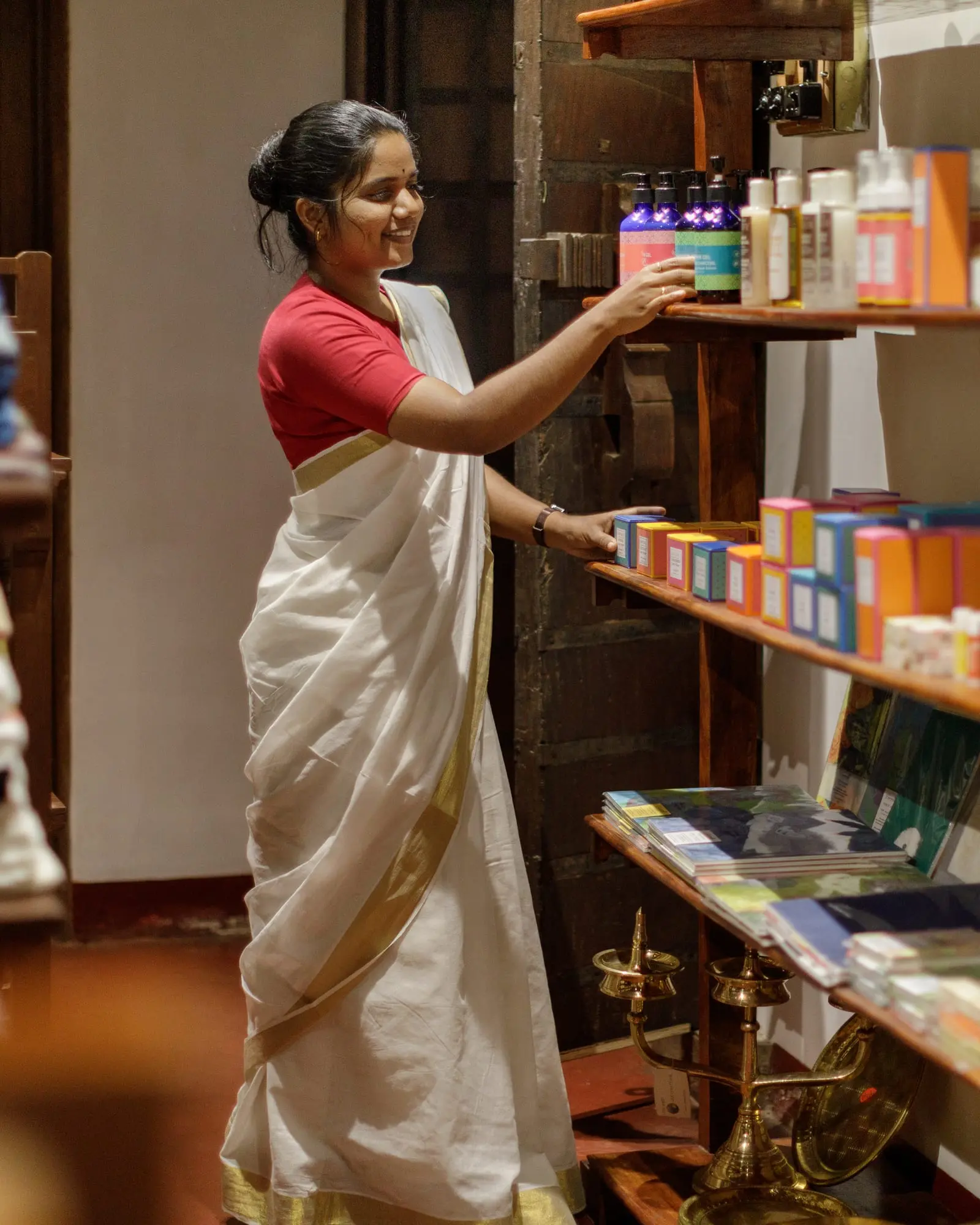 A practitioner in a cream sari arranges oils and preparations on wooden shelves.