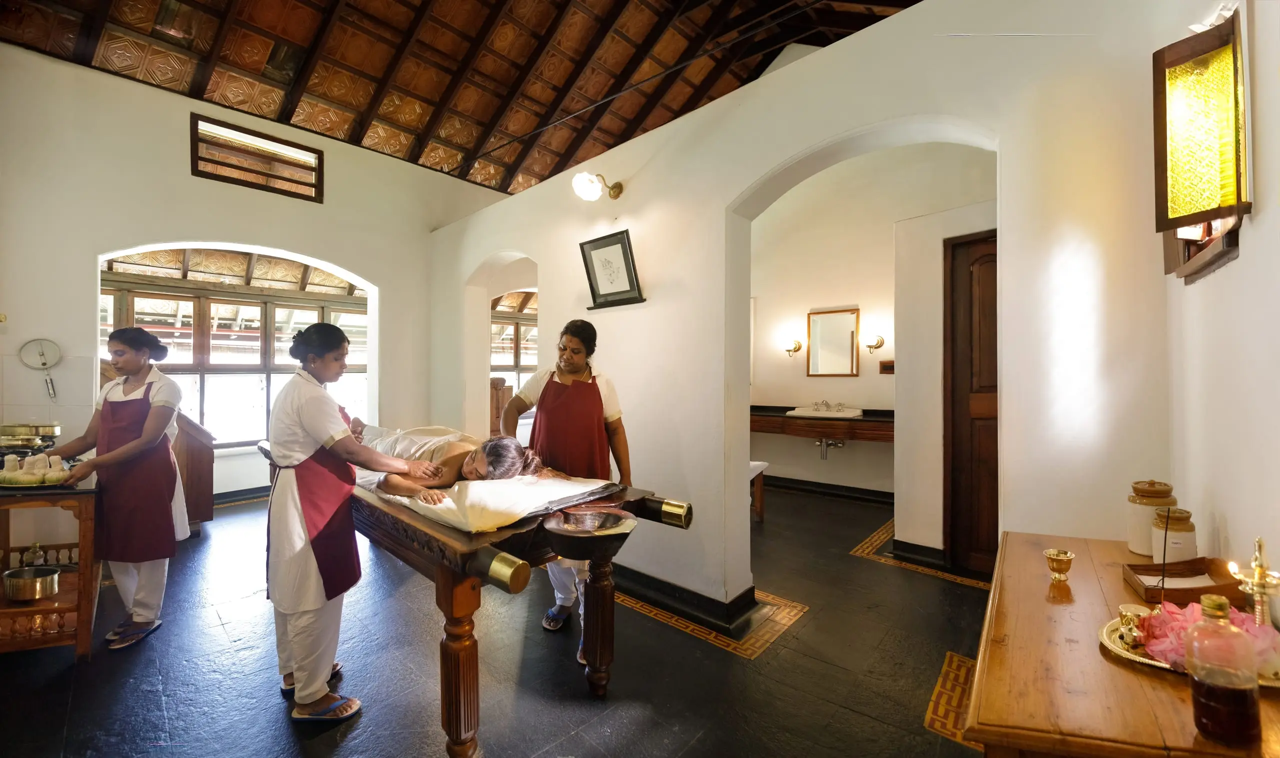 A ritual room of timber and whitewash; practitioners attend a guest at a carved wooden table.