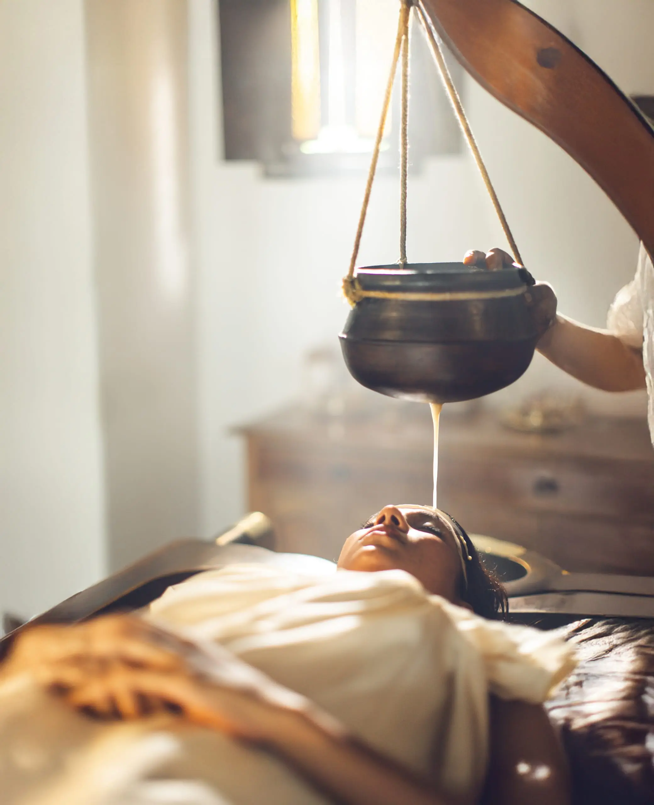 A steady stream of herb-infused milk falls from a hanging vessel to the guest's forehead.