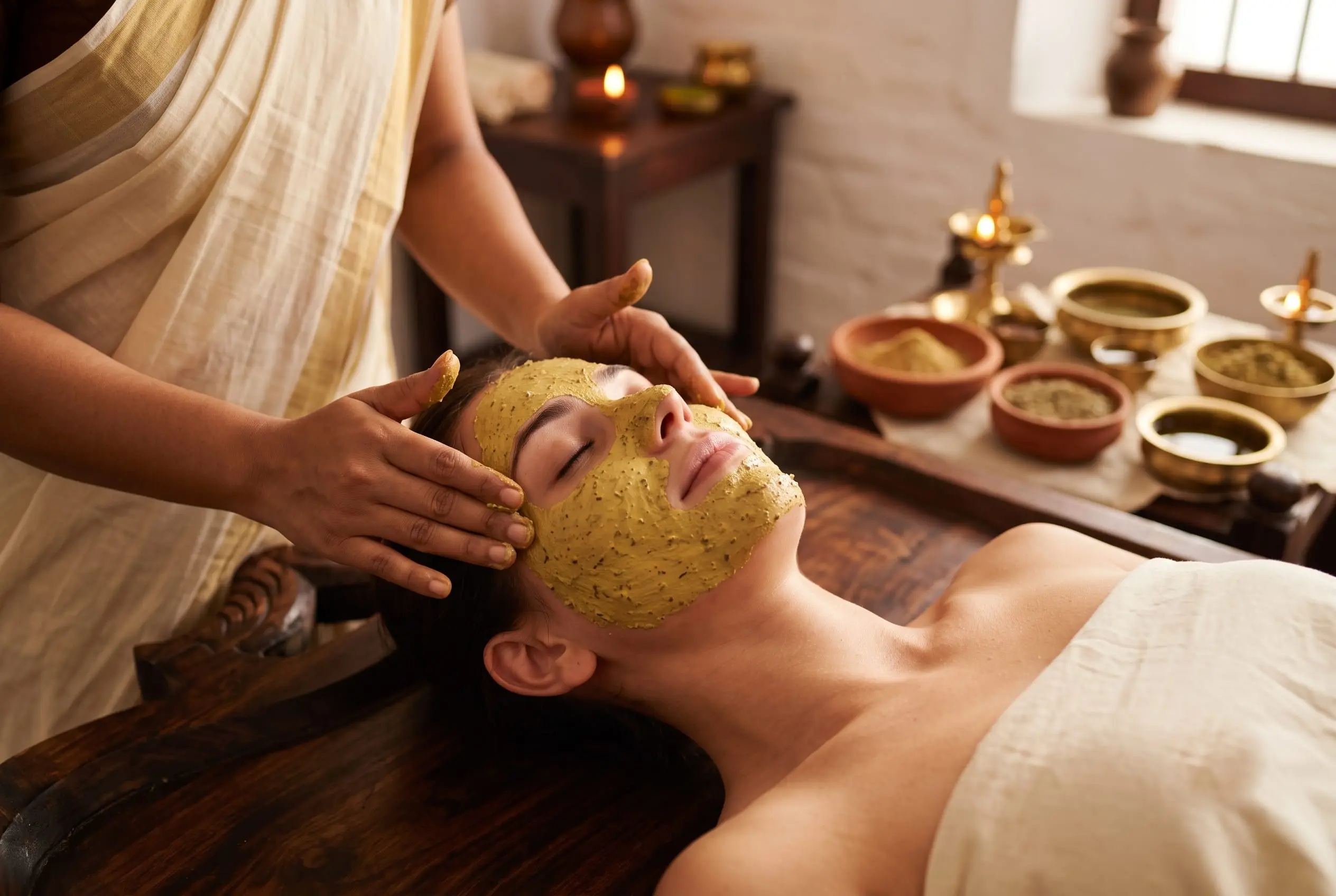 A green herbal pack smoothed over a guest's face; bowls of ground herbs wait beside.