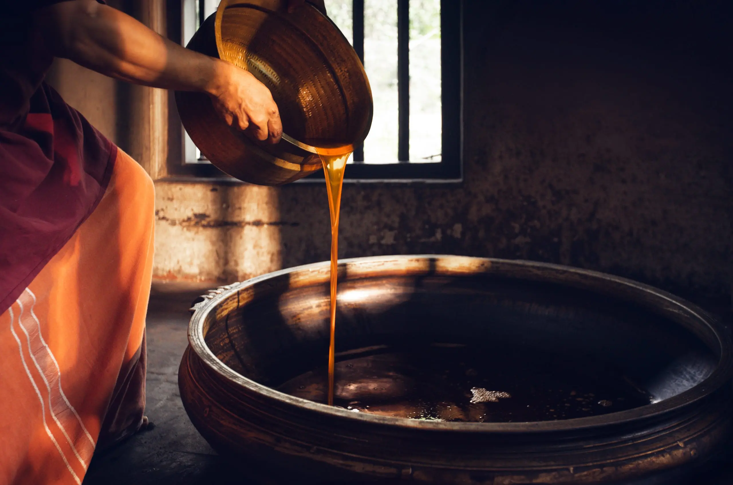 Golden oil poured from a brass basin into a wide iron vessel, by a barred window.