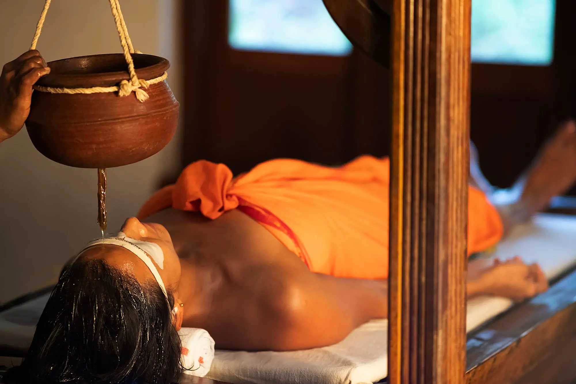 Warm oil falls in a thin stream from a clay vessel to the guest's forehead, eyes covered and closed.