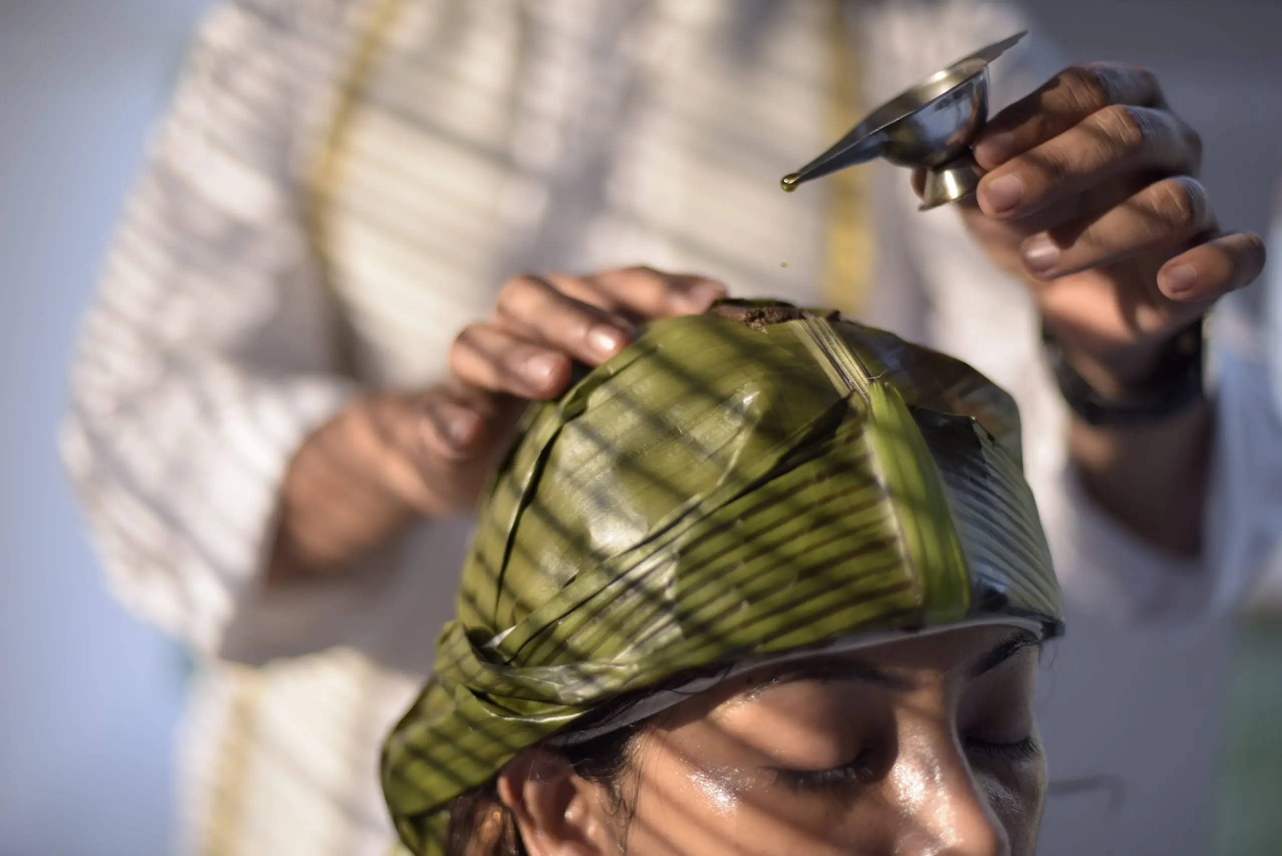A cap of banana leaf holds the herbal pack; oil is dripped slowly onto the crown.